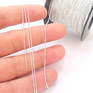 May include: A spool of silver-coloured bead chain. The chain is made of small, round beads that are connected together. The chain is wound around a black spool.