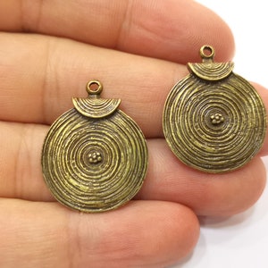 Peut inclure: Deux breloques pendantes en métal doré avec un design circulaire présentant des cercles concentriques et un centre surélevé. Les breloques ont une boucle en haut pour les attacher à des bijoux.