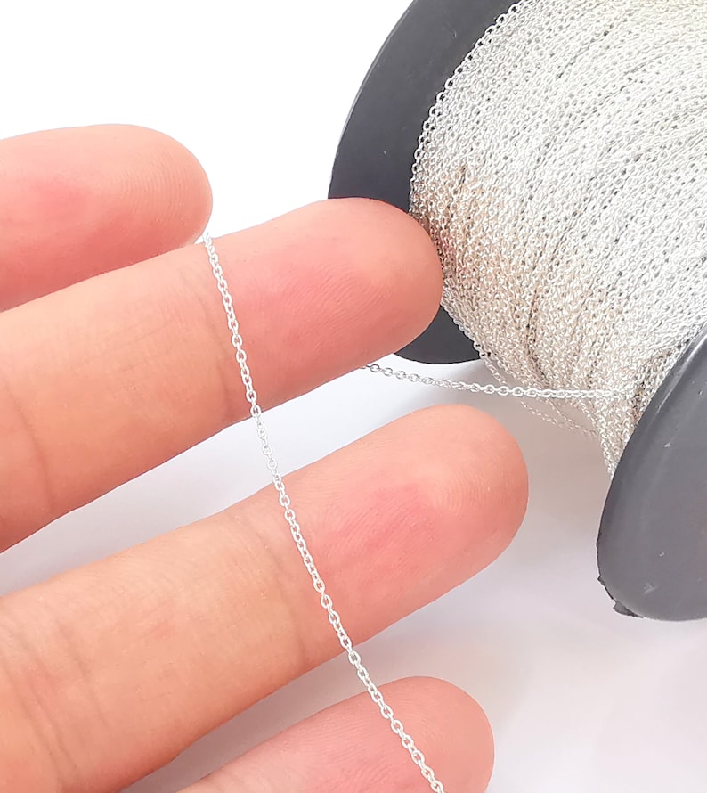 May include: A spool of silver chain, with a length of chain draped over two fingers. The chain is thin and delicate, with a smooth finish.