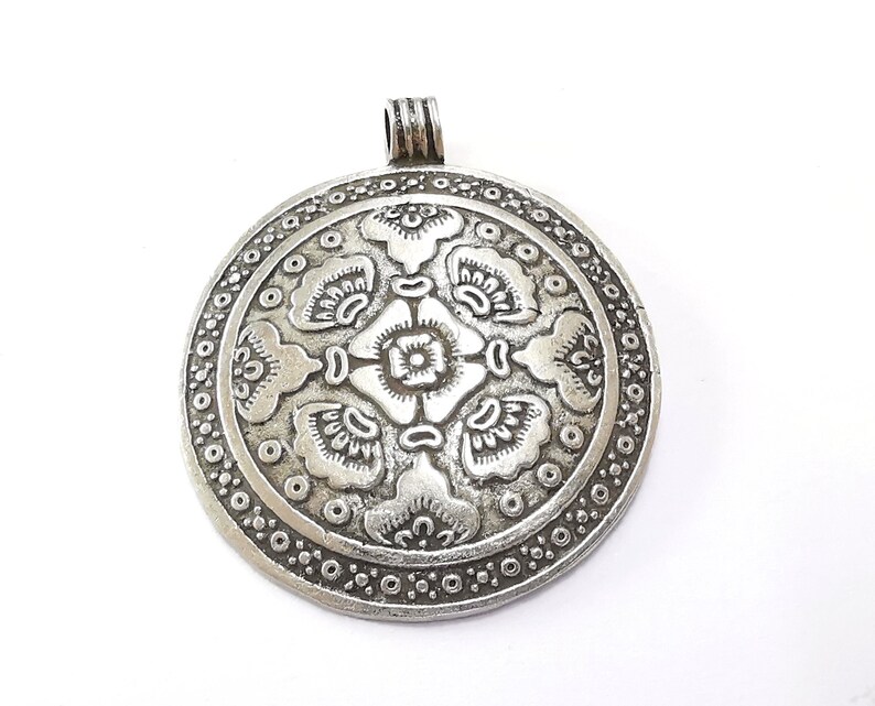 May include: A silver-colored, round pendant with an intricate floral design. The pendant features a central flower surrounded by stylized leaves and a dotted border. It has a loop at the top for a chain.