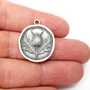 Könnte beinhalten: Silberner Anhänger mit einem detaillierten, geprägten Design einer Distelblüte. Der Anhänger ist rund und hat einen strukturierten Rand.