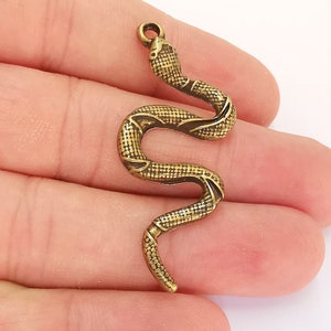 Puede incluir: Un colgante de serpiente de tono dorado con un diseño texturizado detallado. El colgante tiene forma de serpiente con la cabeza levantada y un bucle en la parte superior para sujetarlo a una cadena o cordón.