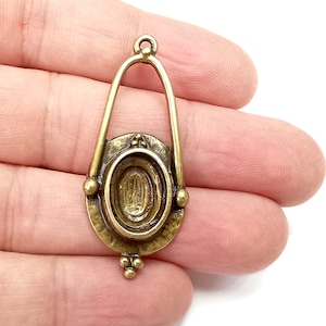 May include: A gold-toned metal pendant with a decorative oval frame and a loop at the top. The pendant has a detailed design with small beads around the oval frame.