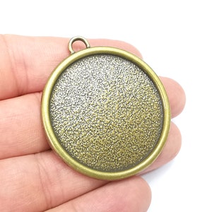 May include: A round, gold-toned metal pendant with a textured surface. The pendant has a loop at the top for attaching a chain or cord.