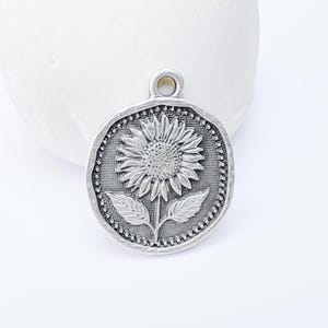 May include: A silver-toned pendant featuring a detailed sunflower design. The pendant has a textured, antique finish and a small loop at the top for hanging. The sunflower is centered on a round, textured background.