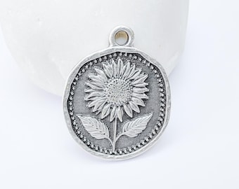 Puede incluir: Colgante plateado con un diseño detallado de girasol. El colgante tiene un acabado texturizado y envejecido y un pequeño lazo en la parte superior para colgarlo. El girasol está centrado en un fondo redondo y texturizado.