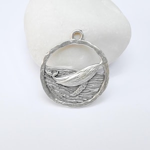 Puede incluir: Encanto de colgante de plata con una ballena saltando fuera del agua. La ballena está en un marco circular con un diseño de agua texturizado.