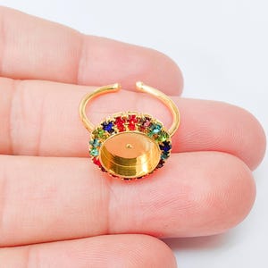 May include: Gold-toned adjustable ring with an oval setting. The ring is adorned with a border of colorful gemstones in red, blue, green, and yellow. The ring's band is open, allowing for size adjustment.