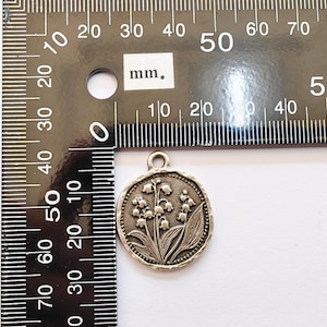 May include: A silver-colored round pendant featuring a detailed relief of lily of the valley flowers and leaves. The pendant is approximately 1 inch in diameter and has a small loop at the top for attaching to a chain or cord. A ruler is in the background.