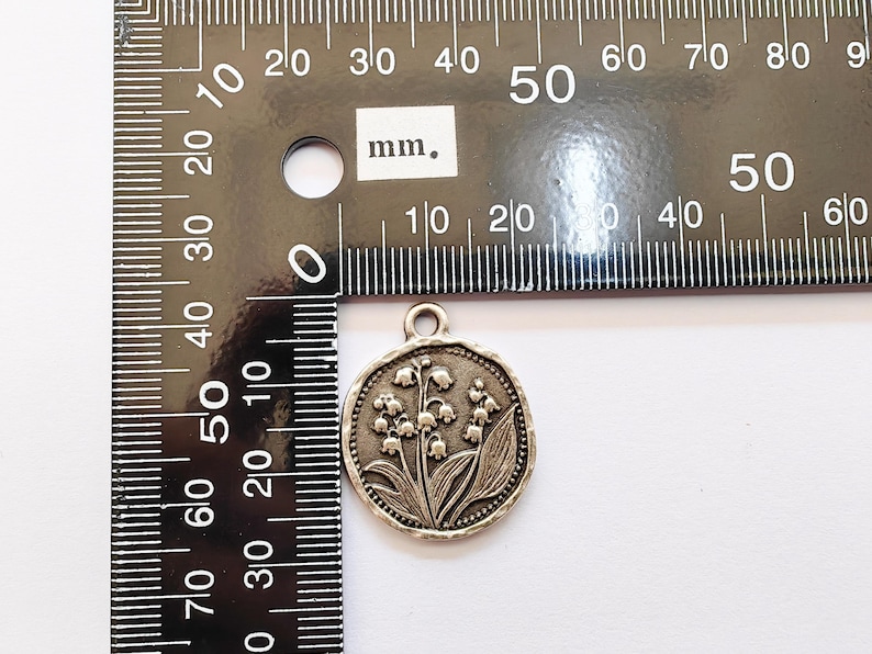 May include: A silver-colored round pendant featuring a detailed relief of lily of the valley flowers and leaves. The pendant is approximately 1 inch in diameter and has a small loop at the top for attaching to a chain or cord. A ruler is in the background.