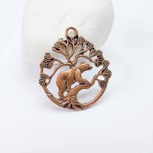 May include: A copper-coloured metal charm featuring a bear standing on a tree branch. The tree is stylised with a circular shape and has a loop at the top for hanging.