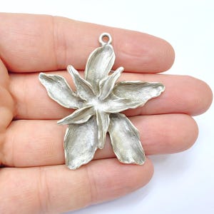 Puede incluir: Un colgante plateado con forma de flor de seis pétalos y un lazo para colgar. El colgante se sostiene en una mano sobre un fondo blanco. La flor tiene una superficie detallada y texturizada.