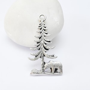 Puede incluir: Encanto de plata con un pino detallado y un oso de pie debajo. El encanto es perfecto para agregar un toque de naturaleza a las joyas.