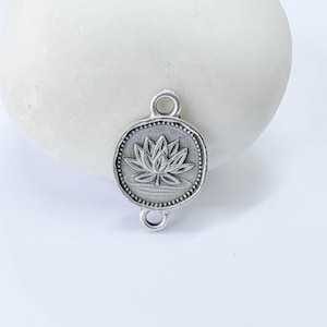 May include: A silver-colored metal charm with a lotus flower design. The charm is rectangular with rounded edges and features a detailed lotus flower in the center, surrounded by a dotted border. It has a loop at the top and a smaller loop at the bottom.
