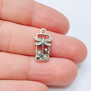 Puede incluir: Un dije rectangular de metal en tono plateado que presenta una libélula detallada con las alas extendidas sobre un grupo de nenúfares. El dije tiene un pequeño lazo circular en la parte superior y un diseño de marco abierto y liso.