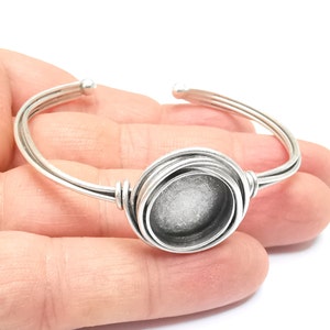 May include: Silver bangle bracelet with a round, silver-toned bezel setting. The bezel is designed to hold a round cabochon or other flat-backed stone.
