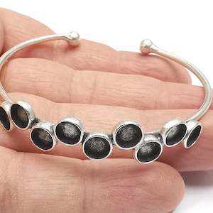 May include: Silver bangle bracelet with a row of nine round, silver charms. The charms have a slightly textured surface and a black center.