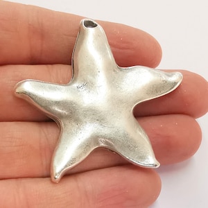 Könnte beinhalten: Ein silberfarbener Metall-Seestern-Anhänger mit einer glatten, leicht strukturierten Oberfläche. Der Anhänger hat oben ein Loch zum Anbringen an einer Halskette oder einem Armband.
