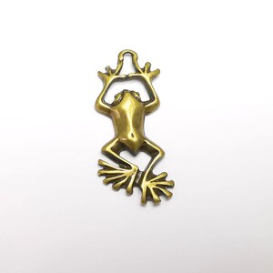 May include: A gold-colored metal frog charm with a hanging loop at the top. The frog is in a sitting position with its legs outstretched and its feet spread out.