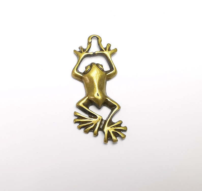 May include: A gold-colored metal frog charm with a hanging loop at the top. The frog is in a sitting position with its legs outstretched and its feet spread out.