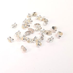 May include: A group of silver earring backs, also known as earring stoppers, scattered on a white surface. The earring backs have a small loop on the back and a flat, round disc on the front.