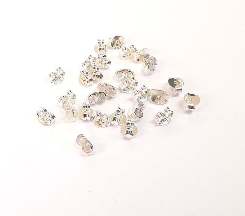 May include: A group of silver earring backs, also known as earring stoppers, scattered on a white surface. The earring backs have a small loop on the back and a flat, round disc on the front.