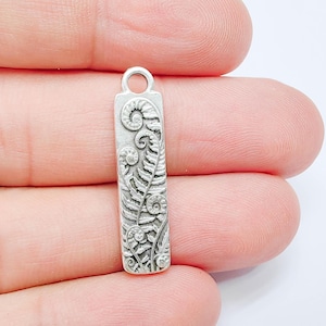 Puede incluir: Un dije rectangular de metal en tono plateado con un patrón de hoja de helecho en relieve y frondas rizadas. El dije tiene un pequeño lazo circular en la parte superior para su fijación, mostrando un diseño botánico detallado y texturizado.