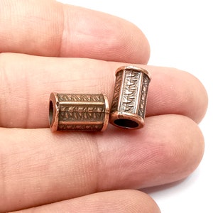 Pode incluir: Duas contas de metal de cor cobre com um padrão geométrico intrincado. As contas são cilíndricas e têm um acabamento liso e polido.