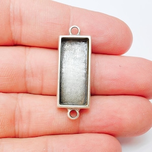 May include: A rectangular silver-toned pendant blank with a recessed area for embellishments. The pendant has two small loops for attaching to a chain or other jewellery components. The blank is held in a hand.