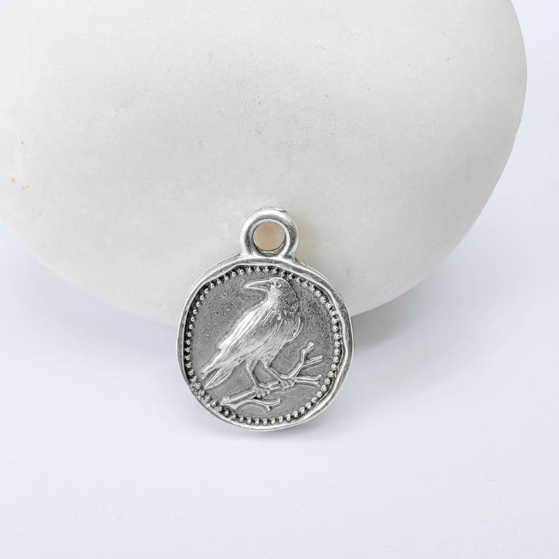 K&ouml;nnte beinhalten: Runder, silberfarbener Anh&auml;nger mit detailliertem Relief einer Kr&auml;he auf einem Ast. Der Anh&auml;nger hat einen strukturierten Rand und eine kleine Schlaufe oben zum Befestigen an einer Kette. Ideal f&uuml;r die Schmuckherstellung.