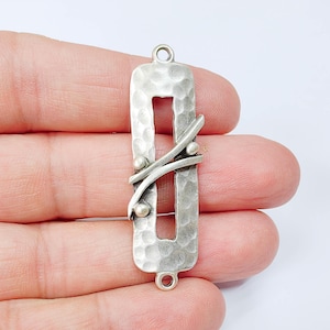 May include: A silver-toned rectangular pendant with a hammered texture and a central cutout. Two curved lines and small spheres are artfully arranged across the surface. The pendant has loops at the top and bottom for attaching to a chain or other jewelry components.