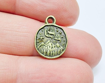 May include: A small, round, antique brass charm featuring a mountain range, trees, and the aurora borealis. The charm has a loop at the top for attaching to a necklace or bracelet. The charm is a detailed, nature-themed pendant.