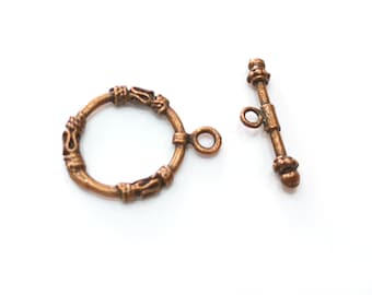May include: A bronze-colored metal clasp with a decorative ring and a toggle bar. The ring has a detailed design and the toggle bar has a small knob at the end.