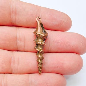 May include: A bronze-colored, spiral-shaped pendant with a loop at the top. The pendant has a textured, detailed design and is held between fingers. This jewelry piece is suitable for necklaces or earrings.