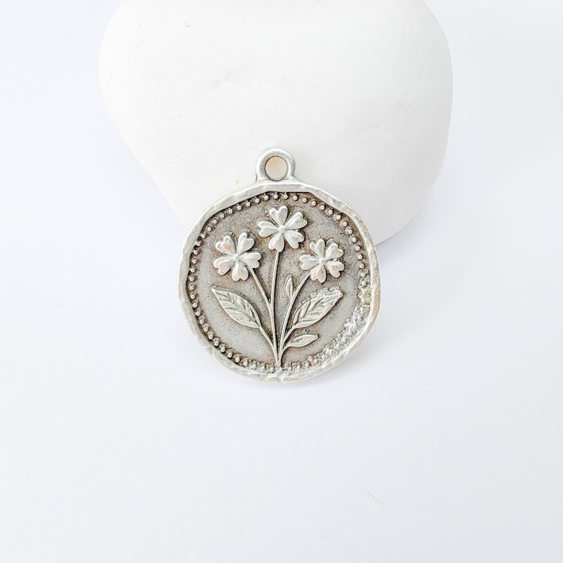 May include: A silver-toned pendant featuring a floral design. The pendant has a circular shape with a beaded border. The central design shows three flowers with leaves and stems. The pendant has a small loop at the top for hanging.