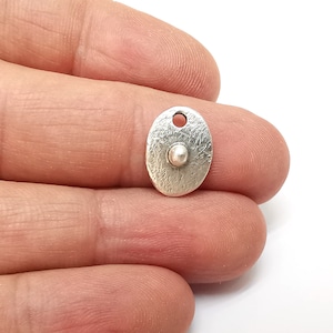 Op de afbeelding: Zilveren ovale bedel met een getextureerd oppervlak en een kleine ronde kraal in het midden. De bedel heeft een gat aan de bovenkant om aan een ketting of armband te bevestigen.