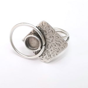 Puede incluir: Un anillo de plata con un bisel cuadrado y una abertura circular. El bisel tiene un diseño texturizado y el anillo está hecho de dos bandas.