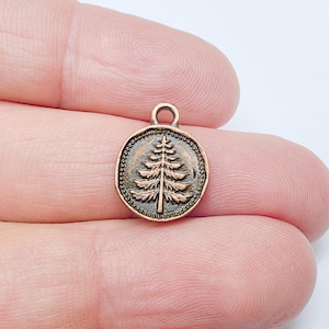 Puede incluir: Un pequeño dije redondo de color cobre con una imagen en relieve detallada de un pino. El dije tiene un lazo en la parte superior para sujetarlo a un collar o pulsera. El diseño del árbol está centrado dentro de un borde de cuentas.