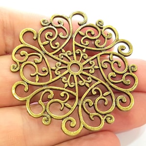 May include: A gold metal filigree charm with a circular design featuring intricate swirls and loops. The charm has a delicate and ornate appearance.