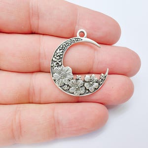 May include: A silver-toned crescent moon charm with floral detailing. The charm features a crescent shape with a loop at the top for hanging. The moon is decorated with detailed flowers and leaves.