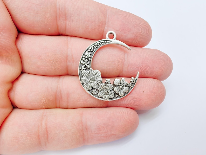 May include: A silver-toned crescent moon charm with floral detailing. The charm features a crescent shape with a loop at the top for hanging. The moon is decorated with detailed flowers and leaves.