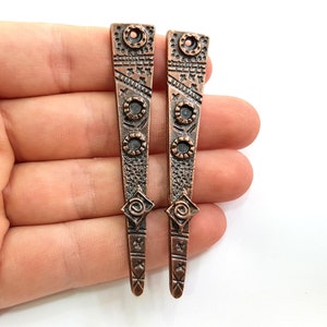 Puede incluir: Dos colgantes de metal de color cobre con intrincados diseños geométricos. Cada colgante tiene un pequeño agujero en la parte superior para colgar.