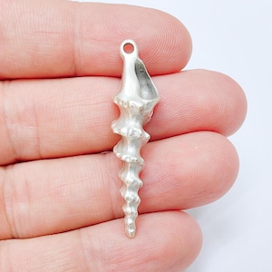 Puede incluir: Un colgante plateado en forma de espiral con un lazo en la parte superior. El colgante tiene un diseño texturizado y acanalado y una parte inferior puntiaguda. La forma general se asemeja a una concha marina o cuerno estilizado. El colgante está en la mano de una persona.