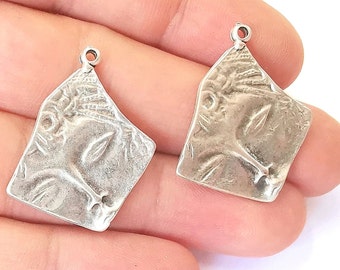 May include: Two silver pendant charms with a detailed face design. The charms are in the shape of a square with rounded corners.