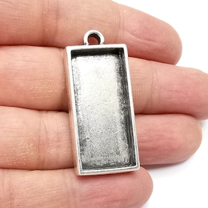 May include: A rectangular, silver-toned pendant tray with a loop at the top for attaching to a chain. The tray has a recessed area for setting a design or image. The pendant is held in a hand.