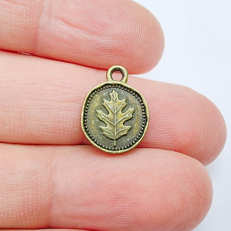 May include: A small, antique brass-colored charm with a detailed oak leaf design. The charm is round with a beaded border and a small loop at the top for attaching to a necklace or bracelet. The leaf is the focal point.