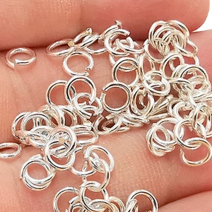 May include: A pile of silver jump rings, a type of jewellery finding, are shown on a hand. The jump rings are small and round, and they are perfect for connecting different pieces of jewellery together.