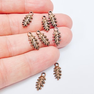 Puede incluir: Una colección de pequeños dijes de hojas de color cobre. Cada dije presenta un diseño detallado de hoja con un pequeño lazo en la parte superior para sujetarlo a joyas u otras manualidades. Los dijes están dispuestos sobre un fondo claro.