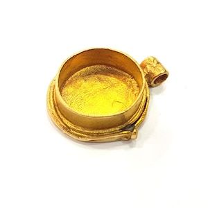 Könnte beinhalten: Ein goldfarbener Anhänger mit einer runden, offenen Oberseite und einer kleinen Schlaufe an der Seite. Der Anhänger ist so konzipiert, dass er ein kleines Objekt, wie ein Foto oder ein Schmuckstück, aufnehmen kann.