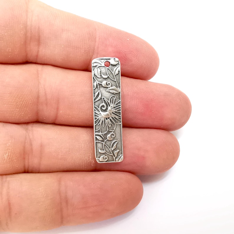 Pu&ograve; includere: Ciondolo in argento con un design floreale. Il ciondolo &egrave; rettangolare e ha un foro nella parte superiore per essere attaccato a una collana o un braccialetto.